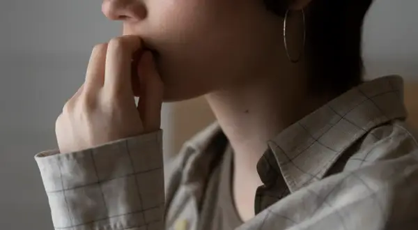 En person med fingrarna vid munnen, en stor ringörhänge syns. Känslan av avslappning eller stress kan anas i den stilla gesten.