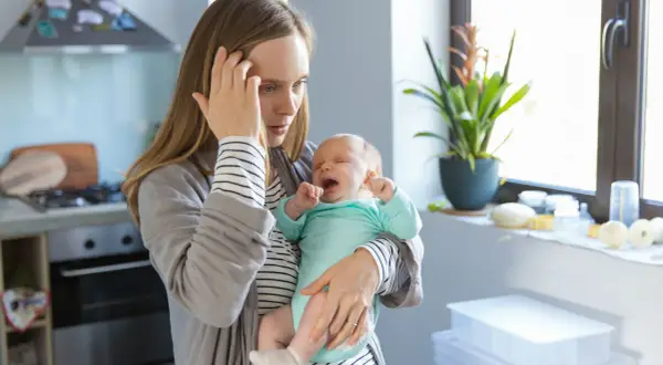 Erschöpfte Mutter hält weinendes Baby. Der 12 wochen schub macht ihr zu schaffen. Sie hält sich die Stirn.