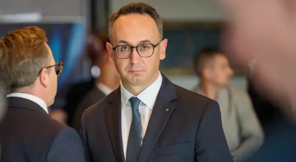 Minister infrastruktury w okularach i garniturze, z zieloną koniczynką na klapie marynarki, rozmawia z kimś na tle innych osób.