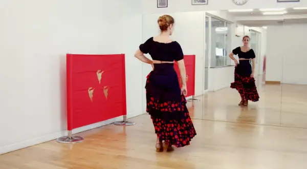 Bailarina con falda flamenca y top negro, practicando una rumbita flamenca frente a un espejo y paneles rojos con detalles dorados.