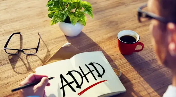 Osoba zmagająca się ze spektrum ADHD pisze w zeszycie. Obok leżą okulary, filiżanka kawy i roślina.