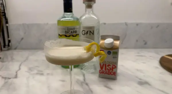 En krämig limoncello drink med citronskal, bredvid flaskor med limoncello, gin och vispgrädde.