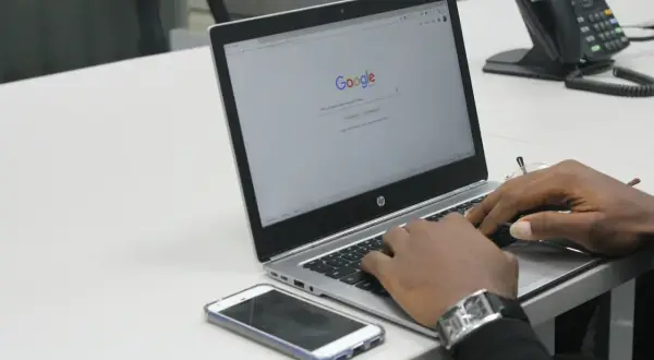 Dłonie piszące na laptopie z otwartą stroną Google, obok leży smartfon.
