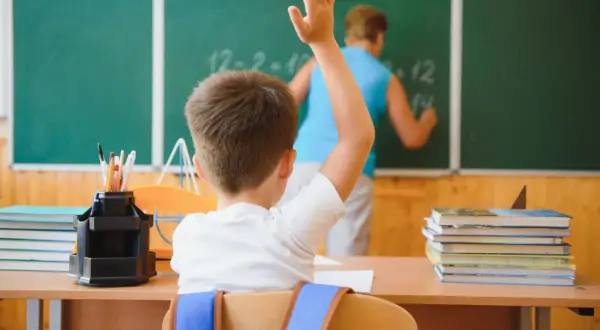 Uczeń z podniesioną ręką przed tablicą, na której nauczyciel pisze.