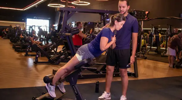 En kvinna utför 45 graders hyperextension med en tränare som ger stöd. Träningspasset sker på ett modernt gym.