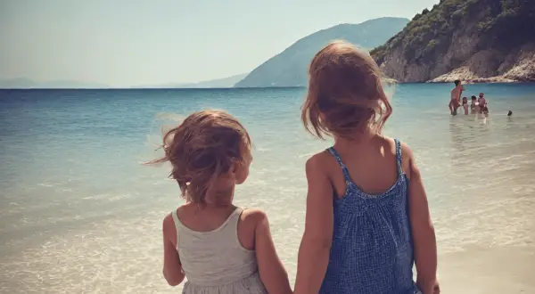 Dwie dziewczynki trzymają się za ręce na plaży, patrząc na morze. W tle widać góry i innych ludzi kąpiących się w wodzie. To idealne wakacje w Albanii z dziećmi.