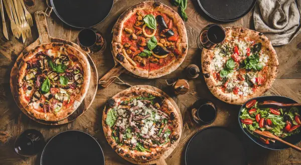 Vier Pizzen, ein Salat und Rotwein auf Holztisch. Die Frage welcher Wein zu Pizza wird hier beantwortet.