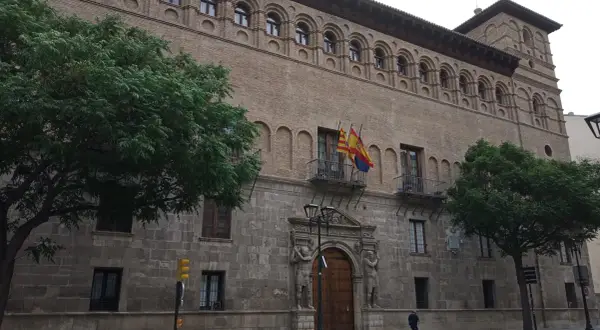 Fachada del palacio de los Luna en Zaragoza, con sus arcos y balcones, flanqueado por árboles verdes.