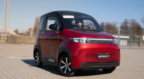 Czerwony, mały samochód elektryczny z kobietą za kierownicą, na tle placu i nieba.