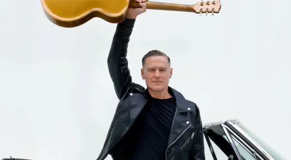 Bryan Adams w skórzanej kurtce unosi gitarę nad głową, gotowy na koncert. Obok klasyczny samochód.