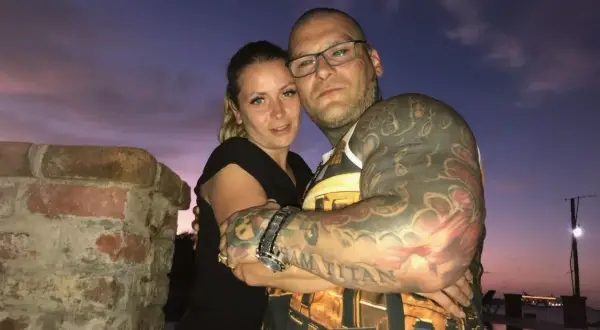 Para, przytulająca się na tle zachodzącego słońca. Ona, jak wygląda żona Popka, ma blond włosy i czarną bluzkę. On, wytatuowany mężczyzna w okularach, obejmuje ją.