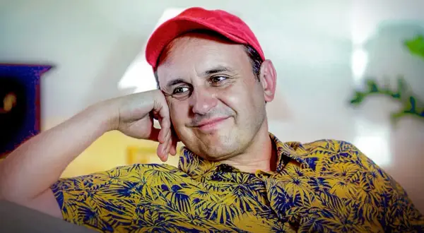 Uśmiechnięty Adam Woronowicz w czerwonej czapce i żółtej koszuli w liście. Wiele jego filmów zapisało się w pamięci widzów.