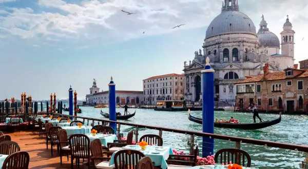 Tavoli apparecchiati su una terrazza con vista sul Canal Grande, gondole e la Basilica di Santa Maria della Salute. Un'ambientazione da sogno per i migliori ristoranti di Venezia.