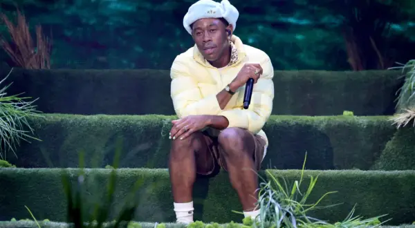 Igor, czyli Tyler, The Creator, siedzi na scenie w żółtej kurtce i jasnoniebieskiej czapce, trzymając mikrofon.