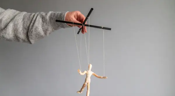 Dłoń w swetrze steruje drewnianym ludzikiem na sznurkach, symbolizując psychologię manipulacji i kontroli.