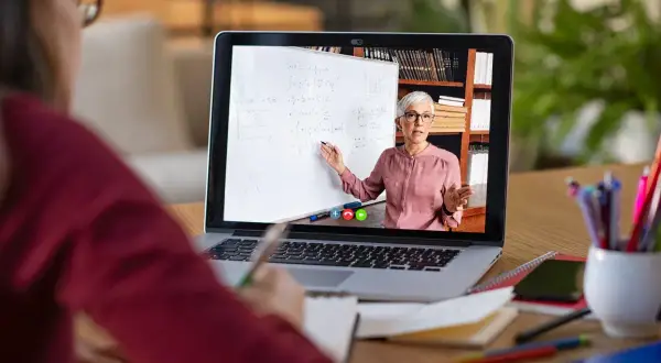 Nauczycielka prowadzi lekcję online na laptopie, wskazując na tablicę z równaniami.