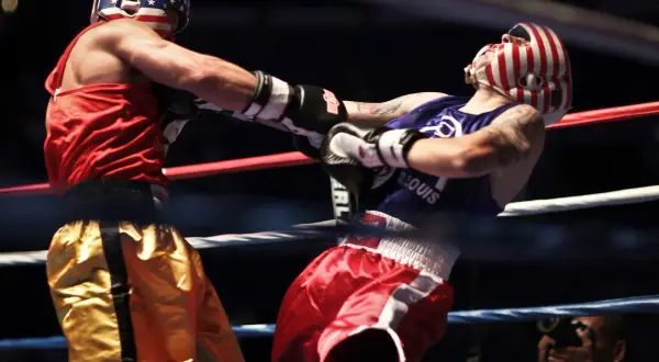 Boxer in red shorts lands a punch to the solar plexus of his opponent in red and white striped headgear.