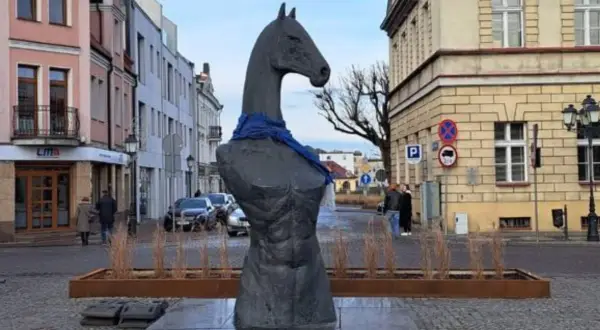 Pomnik z głową konia i niebieską szarfą, przypominający herb. W tle kamienice i ulica.