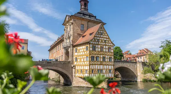 Das Alte Rathaus ragt über den Fluss in der Bamberger Innenstadt. Ein Fachwerkhaus mit gelben Balken und roten Ziegeln schmückt die Brücke.