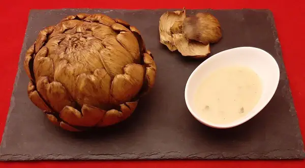 Un artichaut entier, cuit à la cocotte minute, servi avec sa sauce et quelques feuilles détachées sur une ardoise.