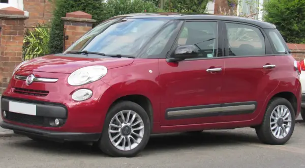 Czerwony Fiat 500 L z czarnym dachem zaparkowany przy ceglanym murze i zieleni.