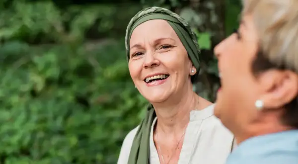 Zwei Frauen lachen im Freien. Die vordere Frau trägt ein Kopftuch und wirkt durch psychoonkologische Beratung gestärkt.