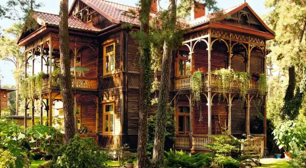 Drewniany, zabytkowy **świdermajer** z werandami i bogatymi zdobieniami, otoczony zielenią i drzewami.