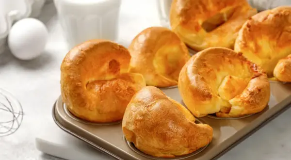 Goldbraune Yorkshire Pudding im Muffinblech, bereit für ein köstliches Rezept. Daneben ein Ei und ein Glas Milch.