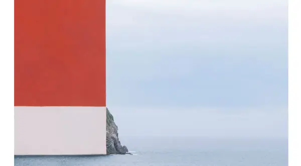 Scogliera rocciosa con mare calmo e cielo azzurro, con un blocco rosso e bianco che incombe a sinistra. Opera di Alberto Selvestrel.