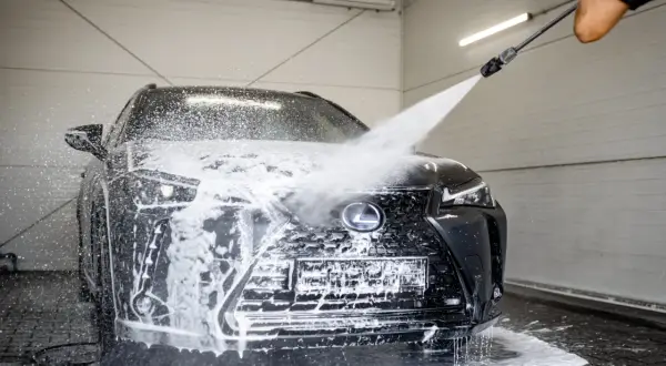 Czarny Lexus jest obficie spieniony podczas mycia na myjni. Widać strumień wody pod ciśnieniem, który spływa po masce i grillu.