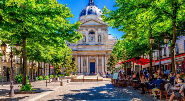 En solig paris april med gröna träd som ramar in en vacker byggnad. Folk sitter vid cafébord i skuggan.