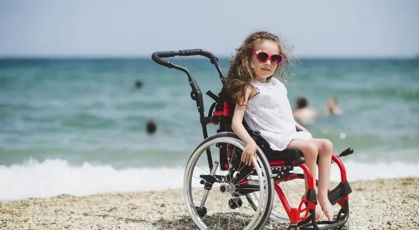 Dziewczynka w okularach przeciwsłonecznych siedzi na plaży w wózku inwalidzkim. W tle widać morze i ludzi kąpiących się.