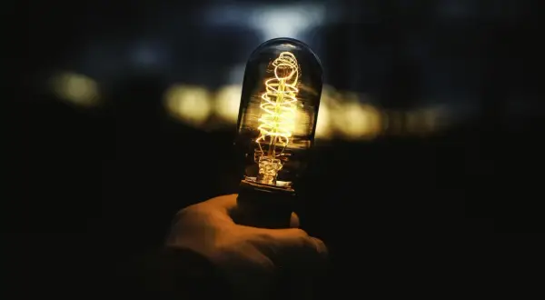 Dłoń trzyma żarówkę 1500 lumenów, której żarnik świeci ciepłym, żółtym światłem. Tło jest ciemne, z rozmytymi, jasnymi plamami.