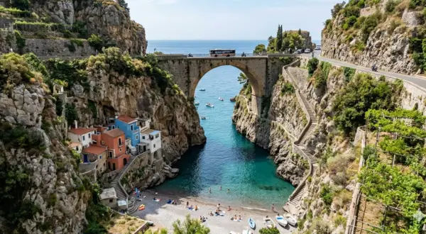 Un ponte ad arco domina un fiordo con case colorate aggrappate alla roccia. Il mare turchese invita a un bagno, mentre la strada panoramica, teatro del furore costiera amalfitana, accoglie bus e moto.