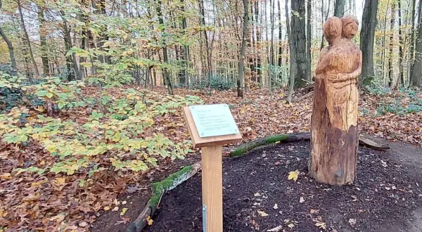 Holzschnitzerei zweier sich umarmender Figuren neben einem Schild mit der Aufschrift "Fatima Kapelle" im Herbstwald.