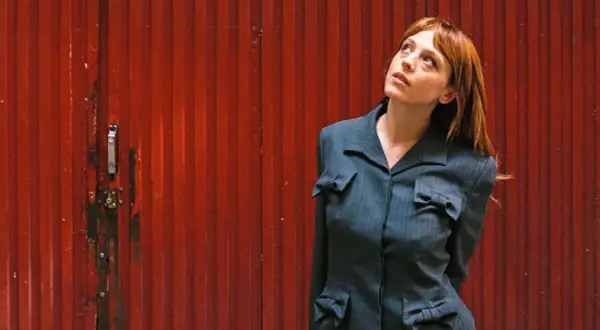 Catalina Sopelana, con su cabello rojizo y mirada perdida, posa frente a una puerta roja acanalada, luciendo un traje gris con detalles de lazos.