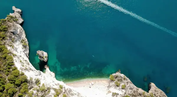 Vista aerea della cala della pipa, con scogliere bianche, vegetazione verde e mare turchese. Una barca lascia una scia sull'acqua.
