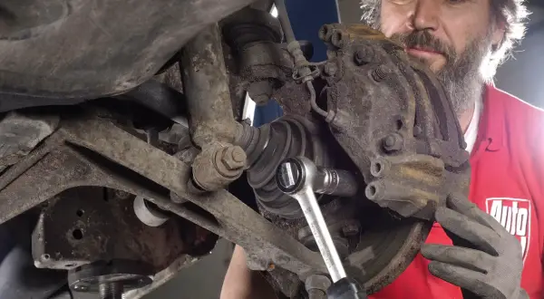 Mechanik w czerwonej koszulce z logo "Auto" dokonuje czyszczenia zacisków hamulcowych, używając klucza nasadowego.