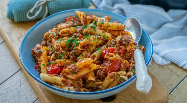 Herzhafte Nudeln mit Hackfleisch und Tomaten, garniert mit frischer Petersilie, serviert in einer blauen Schüssel auf einem Holzbrett.