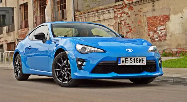 Niebieskie coupe Toyota GR86 z czarnymi felgami stoi przed zniszczoną ceglaną ścianą.