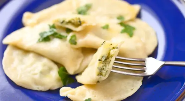 Pyszne pierogi ze szpinakiem i serem, udekorowane natką pietruszki, na niebieskim talerzu. Jedna pierożka jest nadziana widoczna na widelcu.