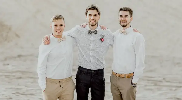 Trzech młodych mężczyzn w eleganckich koszulach i muszkach, gotowych do spełnienia obowiązków świadka na weselu.