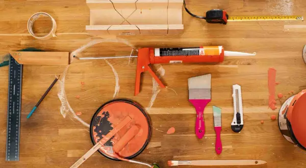 Pistolet à mastic, rouleau, pinceaux et pot de peinture orange sur un établi en bois.