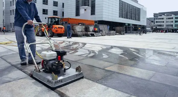 Uomo pulisce autobloccanti con idropulitrice in una piazza. L'acqua riflette la luce sui blocchi di pietra.