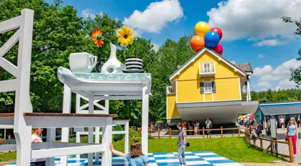Dzieci bawią się w surrealistycznym parku, gdzie dom unosi się na balonach, a stół jest gigantyczny. Idealne miejsce na rodzinne przygody w parkach dla dzieci.