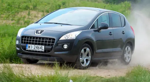 Ciemnoszary Peugeot 3008 2010 na drodze gruntowej, wzbijający tumany kurzu.