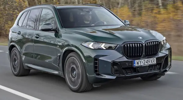 Nowe BMW X5 w ciemnozielonym kolorze mknie po drodze, otoczone jesiennym lasem. Kierowca w okularach pewnie trzyma kierownicę.