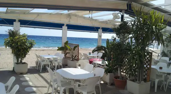 Terraza de restaurante en la playa. Mesas y sillas blancas bajo un toldo, con el mar azul de fondo. Un lugar perfecto para comer con agua amarga.