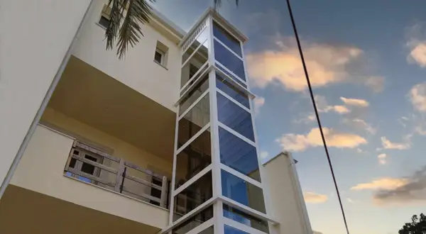 Moderno montacarichi persone esterno in vetro e metallo bianco, che sale lungo un edificio color crema, con cielo nuvoloso al tramonto.