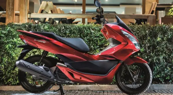 Czerwony skuter Honda PCX, jeden z popularnych **marek skuterów**, zaparkowany przed zielonym żywopłotem.